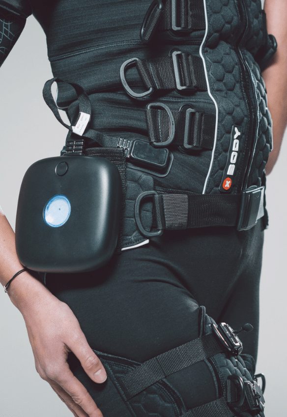 A person wearing a black, high-tech suit with multiple straps and buckles. A small black device with a blue light is attached to the waist, possibly for fitness or medical use.