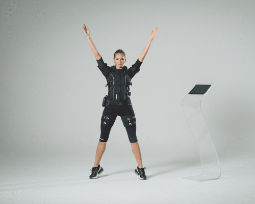 A woman wearing an EMS (electrical muscle stimulation) suit performs a jumping jack with arms raised, facing forward. She stands near a touchscreen device on a clear stand in a minimalist studio setting.