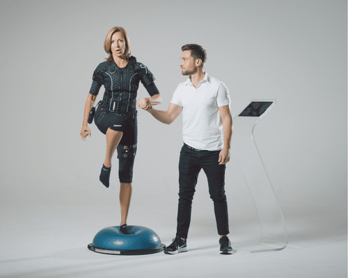 A woman wearing an EMS suit balances on one leg on a BOSU ball while a male trainer in a white shirt and black pants supports her and monitors progress using a nearby digital tablet stand.