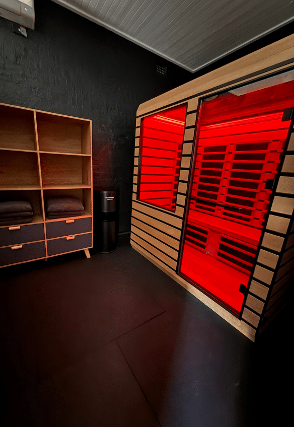 A modern room with black walls features a wooden infrared sauna glowing red, a wooden shelf with dark drawers and folded towels, and a small black water dispenser.