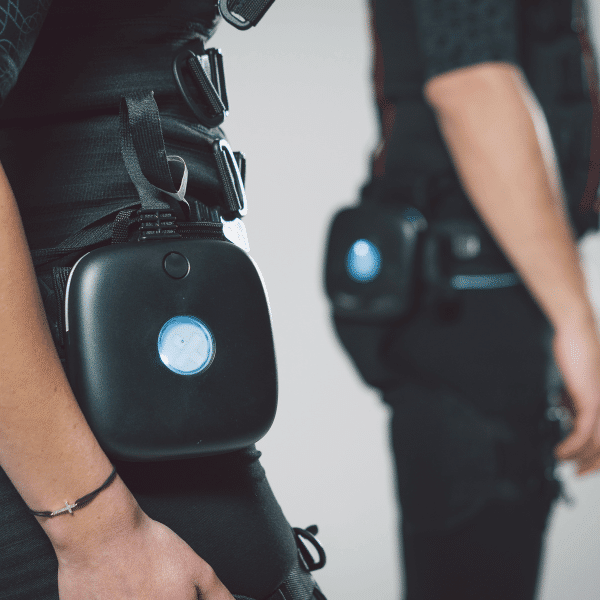 Close-up of two people wearing black suits with attached rectangular black devices featuring a blue circular light, strapped to their waists. The focus is on the device and part of their arms and hands.