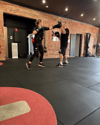 A woman wearing boxing gloves practices punching with a trainer who is holding focus pads in a gym with brick walls and black padded flooring.