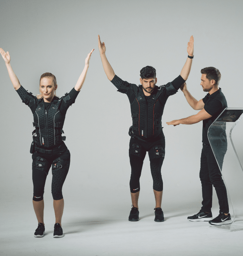 Two people in black EMS training suits stand with arms raised while an instructor, also in black, guides them. The group is indoors with a plain background and a control panel nearby.