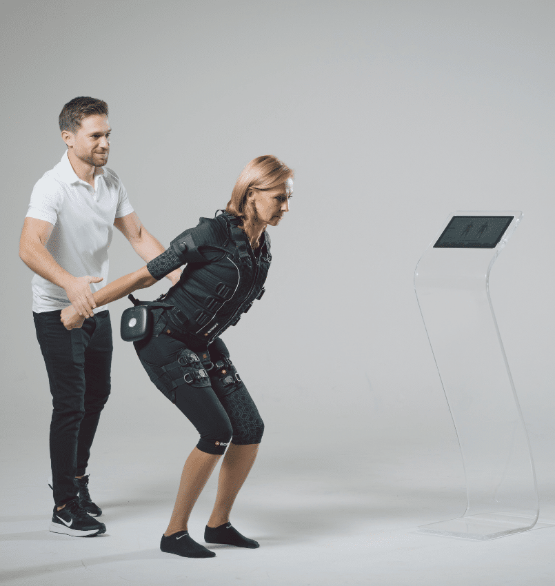 A woman wearing an EMS (electrical muscle stimulation) suit squats with assistance from a man in a white polo shirt. She faces a screen on a clear stand in a bright studio setting.