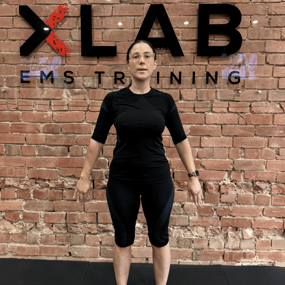 A person in black athletic wear stands facing forward with arms slightly out to the sides. Behind them is a brick wall with the text "XLAB EMS TRAINING" in large black letters.