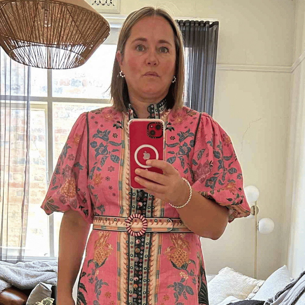 A woman wearing a pink floral dress with puffed sleeves takes a mirror selfie in a bright room with a window, curtains, and a floor lamp in the background.