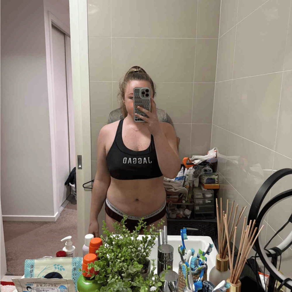 A person in athletic wear takes a mirror selfie in a bathroom. The counter holds plants, toiletries, and toothbrushes. The background shows a partially open door and various items on shelves.