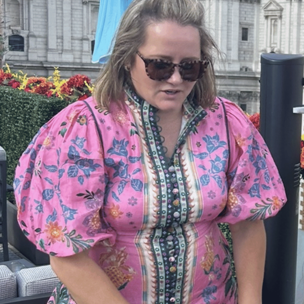 A woman with long blonde hair wearing sunglasses and a pink floral dress with puffed sleeves stands outdoors, with a historic building and colorful flowers in the background.