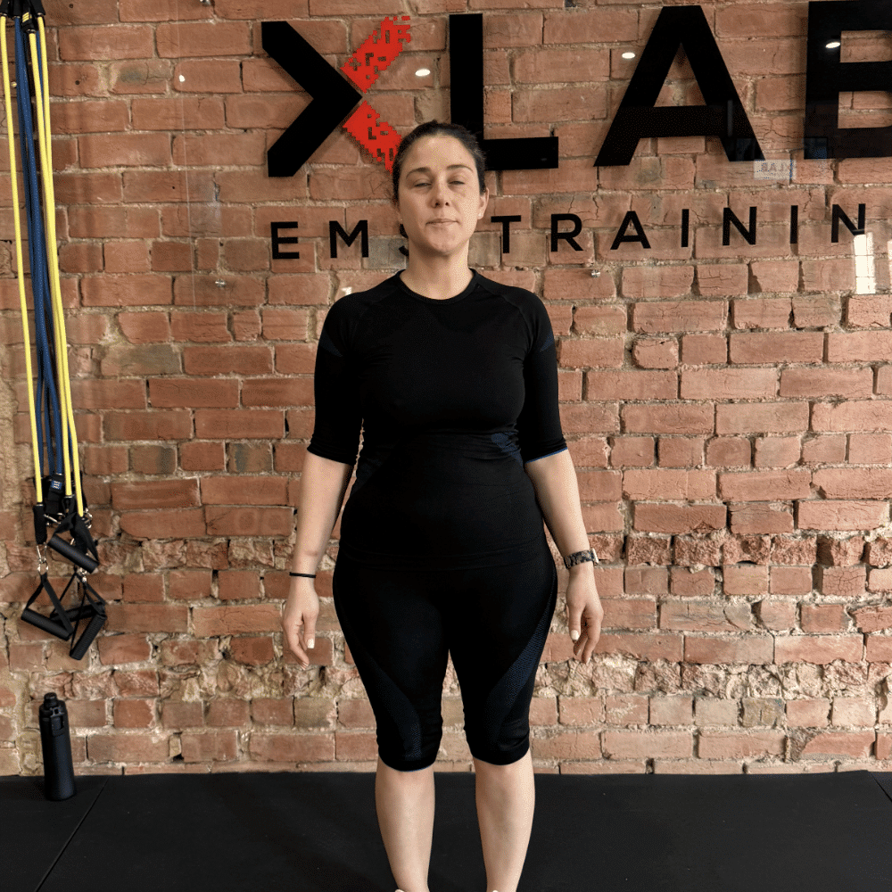 A woman in black athletic wear stands with eyes closed in front of an exposed brick wall with "XLAB EM TRAINING" written on it. Exercise bands hang on the left and a water bottle sits on the floor.