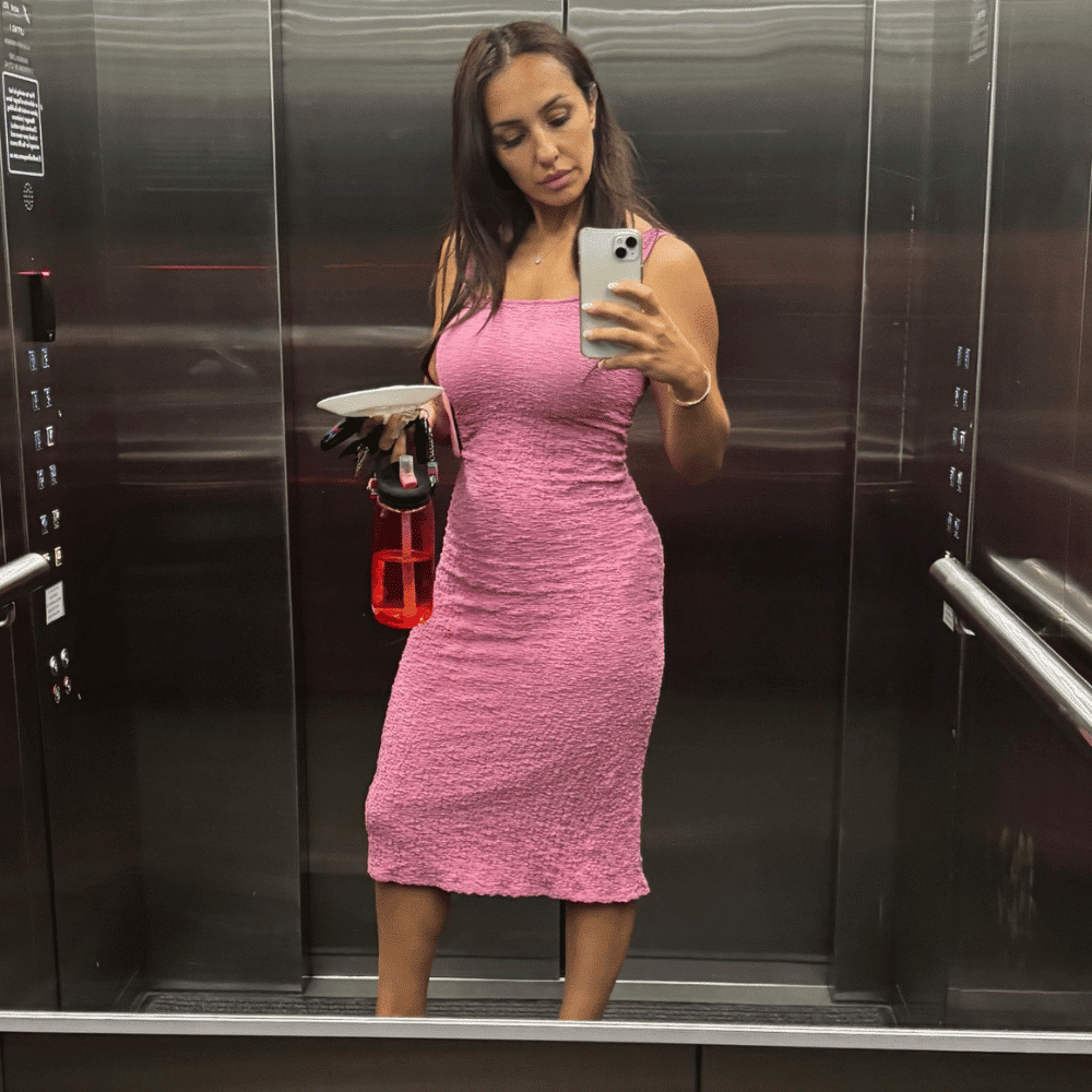 A woman in a fitted pink dress takes a mirror selfie in an elevator while holding a red water bottle and a small plate. The elevator has metallic walls and handrails.