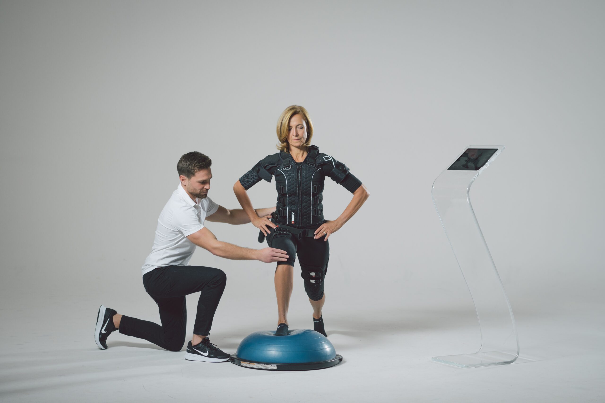 A woman in a black suit stands on a balance trainer while a man in sportswear assists her. They are in a minimal studio with a tablet stand nearby.