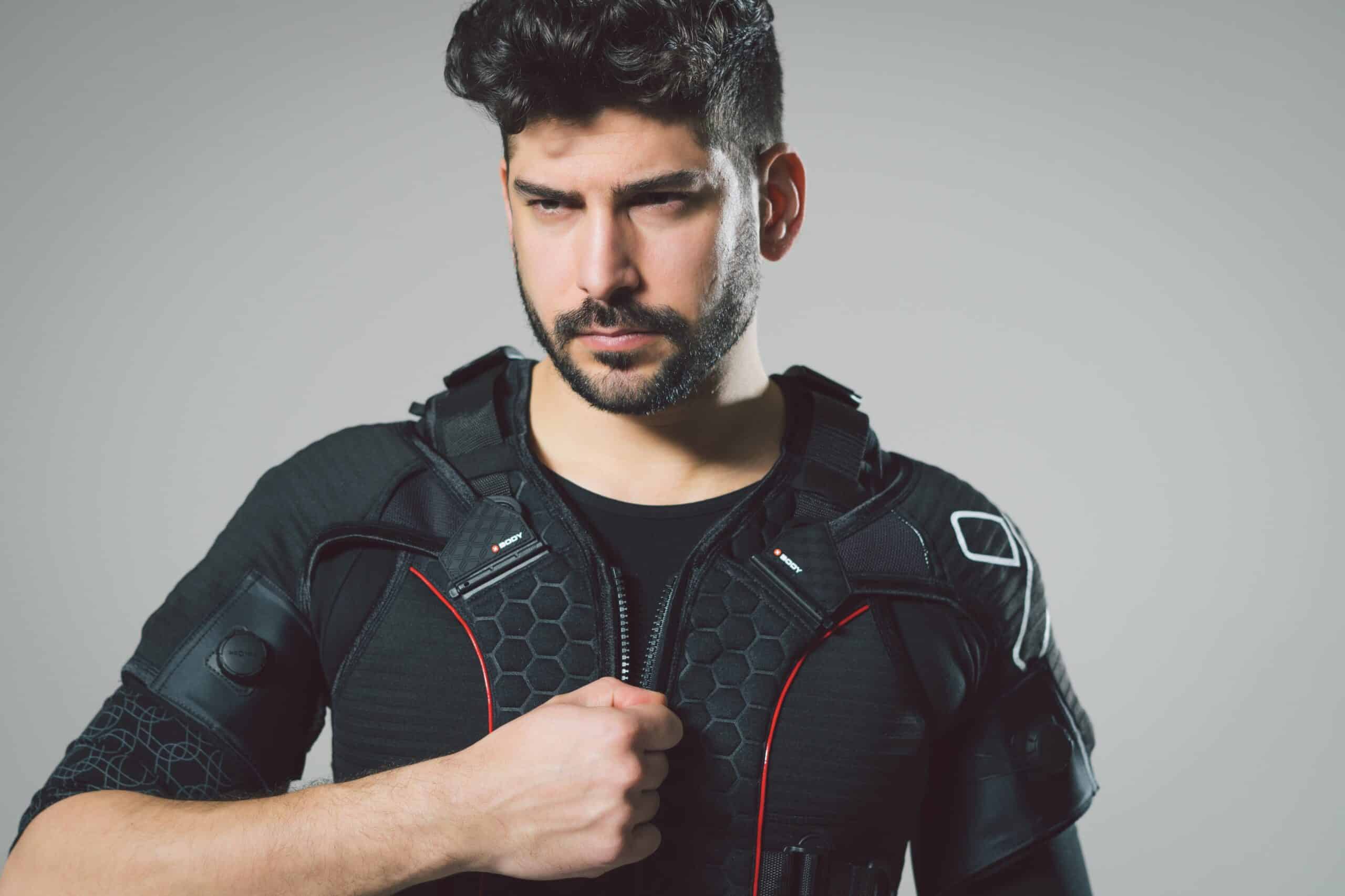 A man with dark hair and a beard wears a futuristic black suit with red accents, gripping the front of the suit with one hand and looking ahead with a focused expression. The background is plain and neutral.