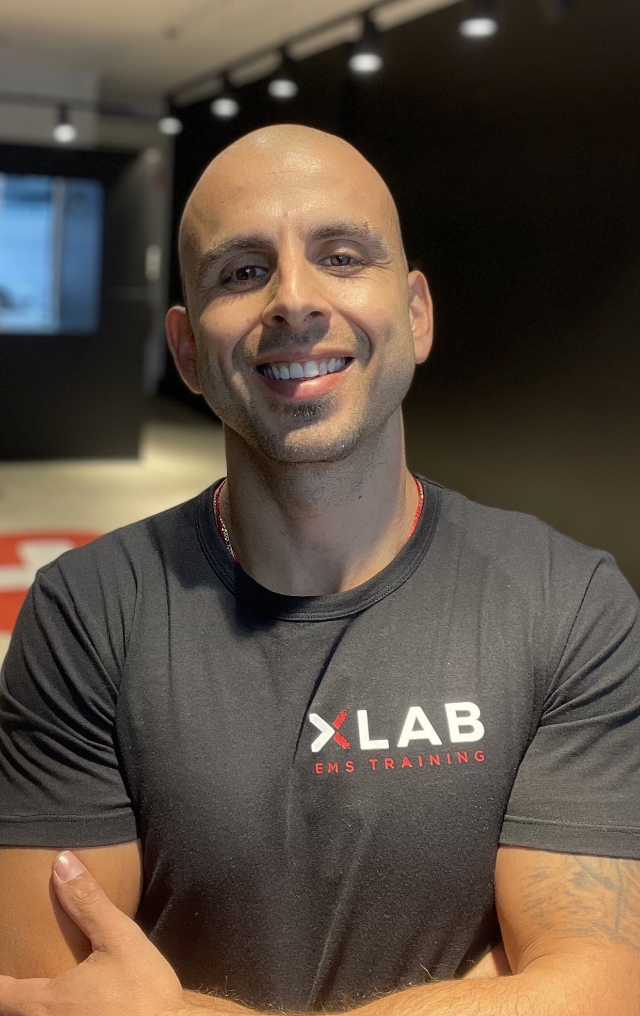 A smiling, bald man with crossed arms wearing a black "XLAB EMS TRAINING" t-shirt stands in a modern gym with dark walls and bright overhead lights.