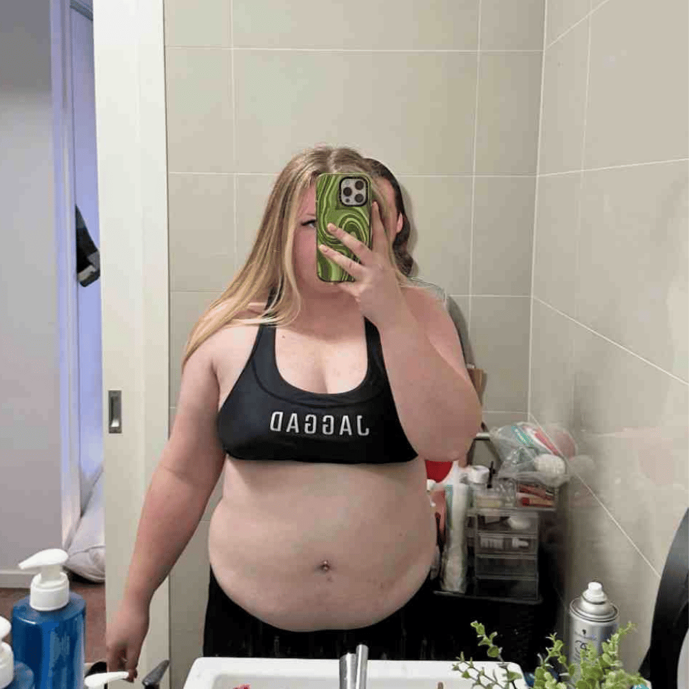 A person with long blonde hair takes a mirror selfie in a bathroom, wearing a black sports bra and holding a green phone. Bathroom items are visible on the counter. The bra text appears reversed in the reflection.