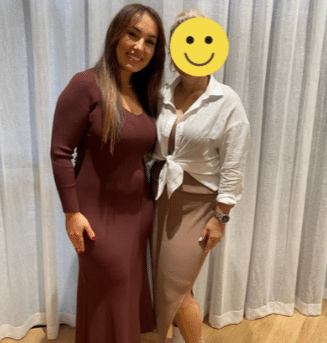 Two women stand side by side indoors. One, in a long-sleeved maroon dress, has long hair and is smiling. The other, whose face is covered with a smiley face, wears a white blouse and beige skirt. Light curtains are behind them.