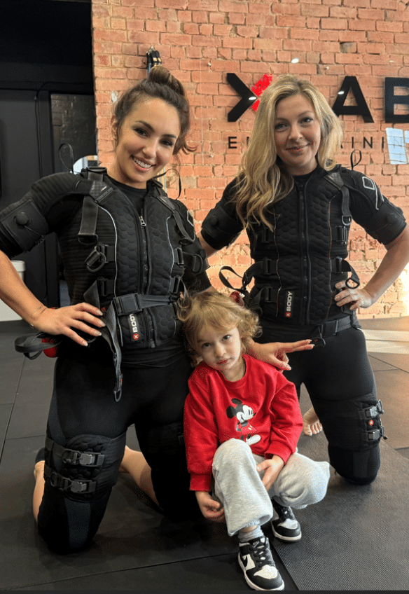 Two women in black EMS training suits kneel on a mat with a young child in a red Mickey Mouse sweatshirt sitting between them. The setting appears to be a fitness studio with a brick wall in the background.